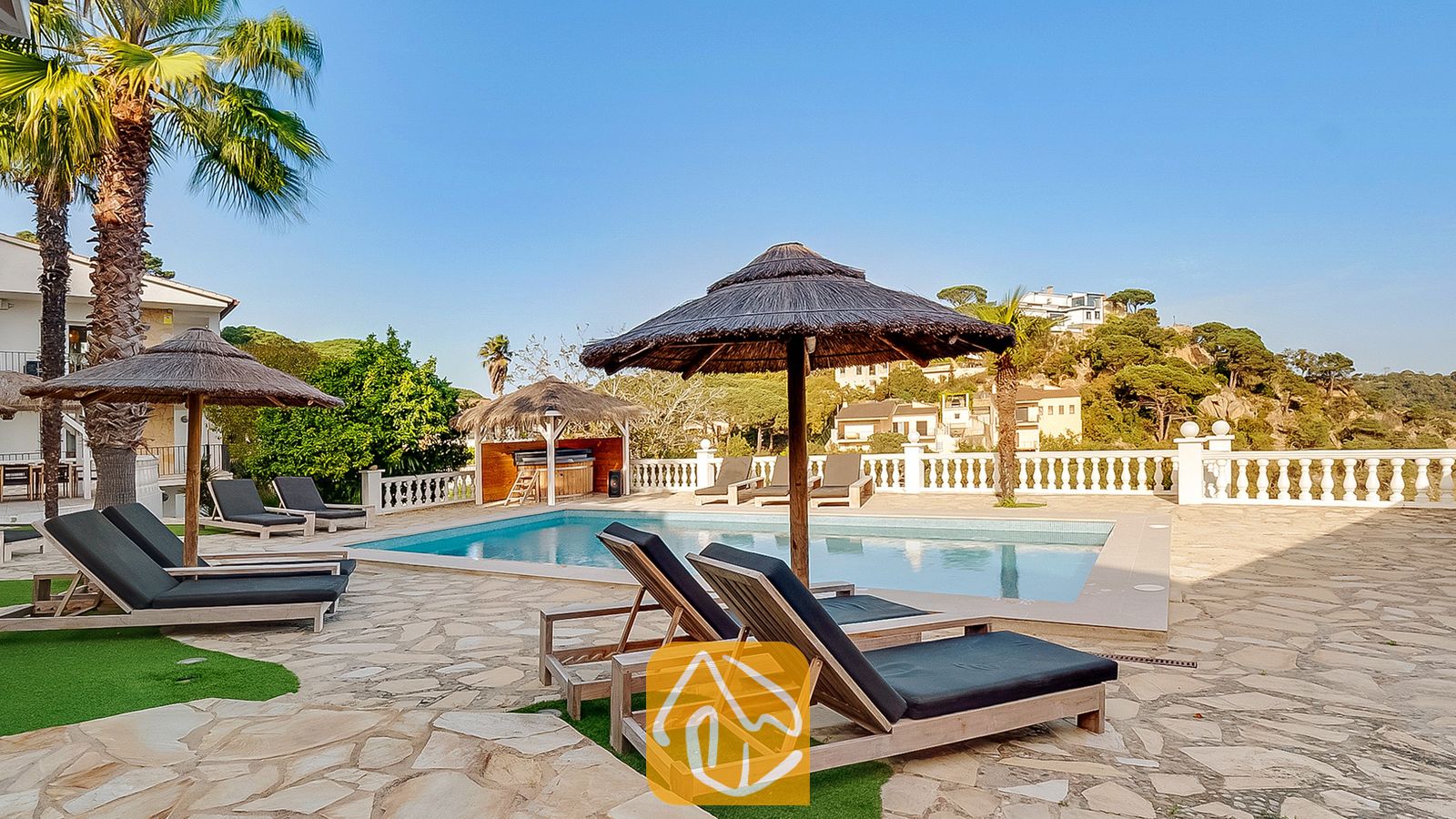 Soleada terraza con piscina en Villa Tropicana, equipada con tumbonas de madera con cojines oscuros, sombrillas de paja y una piscina con el telón de fondo de una colina.