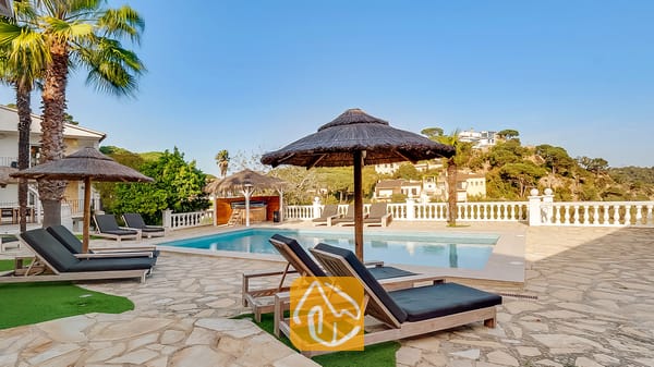 Soleada terraza con piscina en Villa Tropicana, equipada con tumbonas de madera con cojines oscuros, sombrillas de paja y una piscina con el telón de fondo de una colina.
