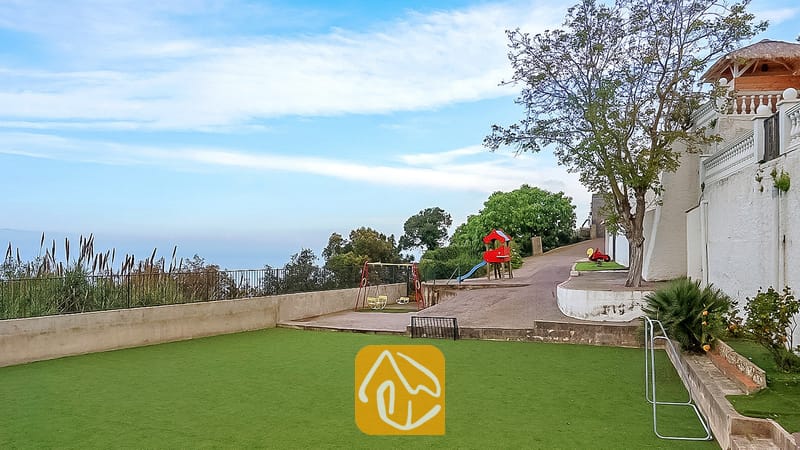 Área de juegos al aire libre en Villa Tropicana que cuenta con un columpio rojo, un tobogán y un césped artificial verde junto a una pared exterior blanca.