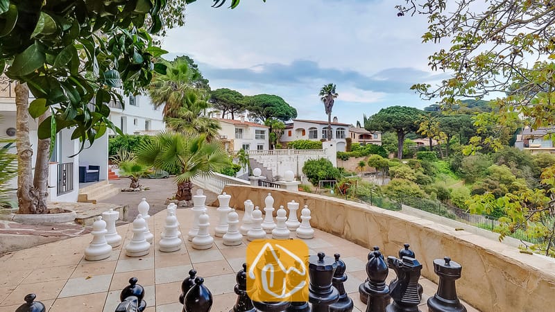 Gigante juego de ajedrez al aire libre en una terraza pavimentada en Villa Tropicana, con vistas a las colinas y árboles españoles circundantes.