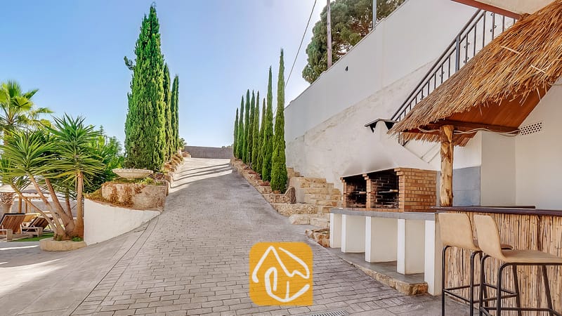 Camino de entrada pavimentado en Villa Tropicana, flanqueado por altos cipreses y equipado con una barbacoa de ladrillo al aire libre y una barra con techo de paja y taburetes altos.