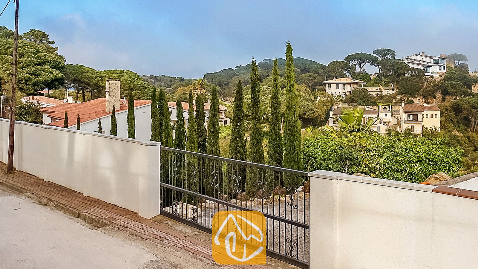Vista exterior de Villa Tropicana que muestra un muro perimetral blanco, un portón de entrada de metal negro y altos cipreses con vistas a un paisaje residencial montañoso.