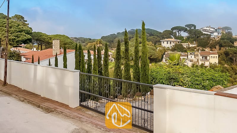 Vista exterior de Villa Tropicana que muestra un muro perimetral blanco, un portón de entrada de metal negro y altos cipreses con vistas a un paisaje residencial montañoso.