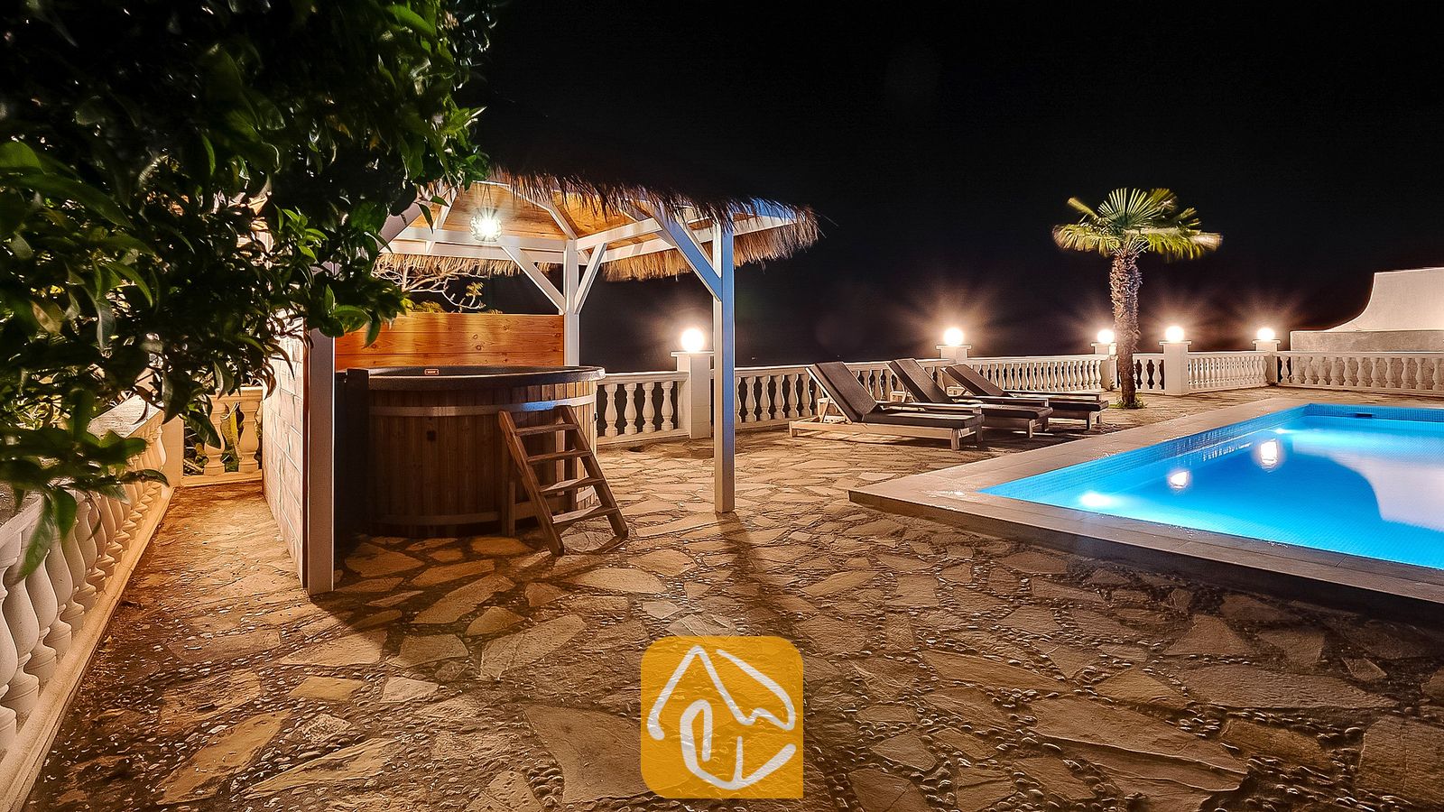 Piscina iluminada y tina de hidromasaje de madera bajo un cenador de paja en el patio de piedra de Villa Tropicana por la noche.
