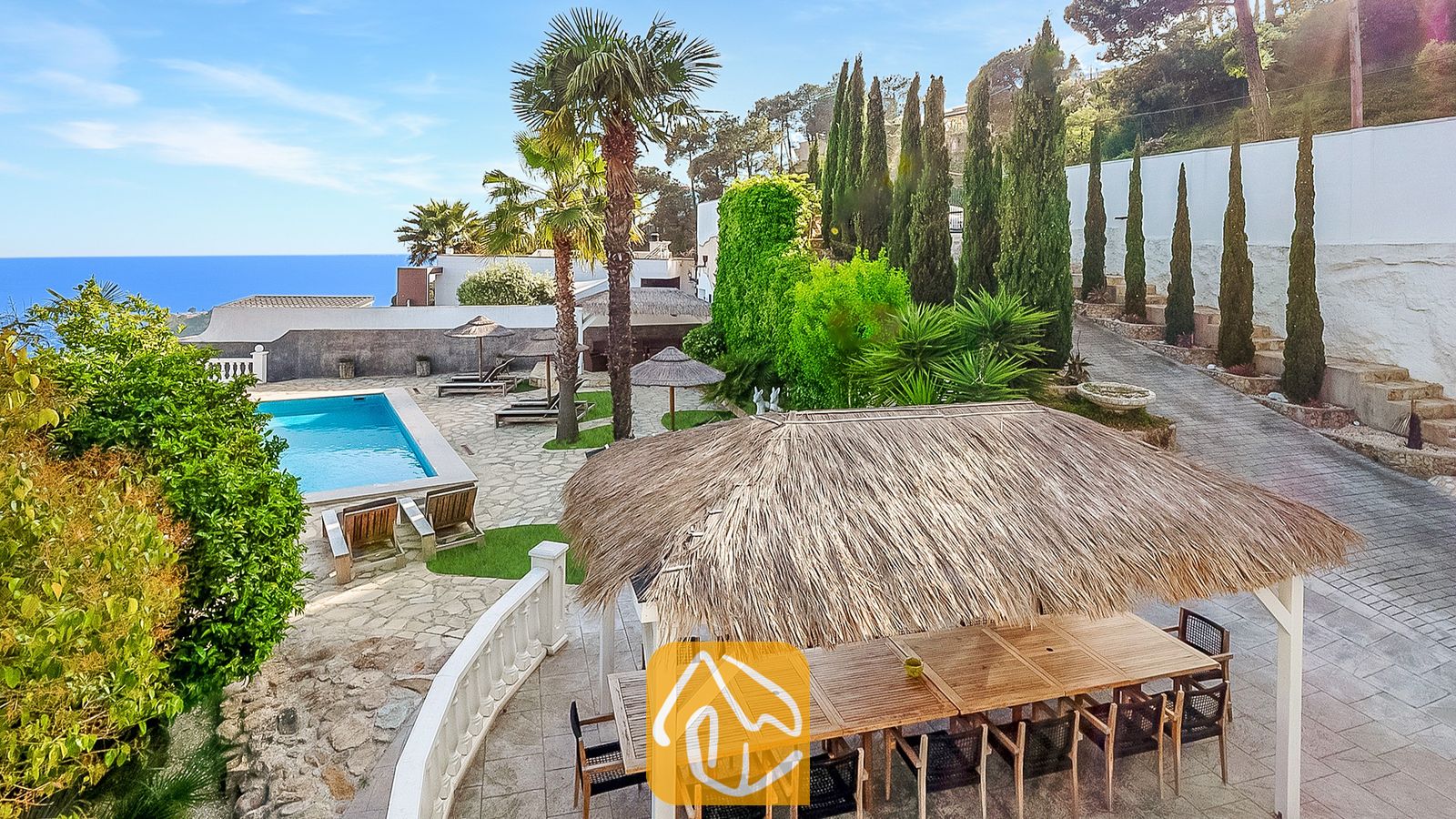 Exterior de Villa Tropicana con una pérgola de comedor con techo de paja, patio de piedra, piscina y vistas al mar bajo un cielo azul.