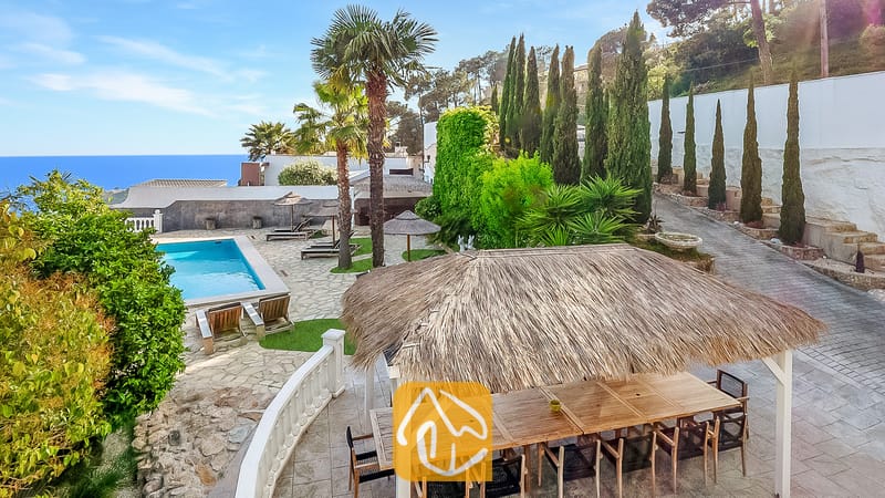 Exterior de Villa Tropicana con una pérgola de comedor con techo de paja, patio de piedra, piscina y vistas al mar bajo un cielo azul.