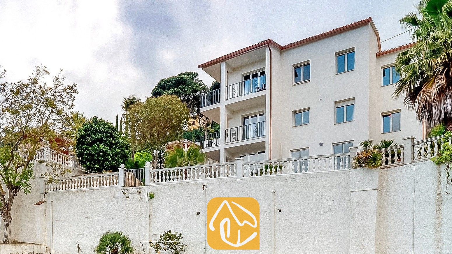 Vista exterior de Villa Tropicana, que muestra un edificio blanco de varias plantas con balcones, tejado de teja de terracota y muros de contención.