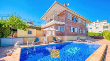 Vista exterior de Villa Beyonce, que cuenta con una piscina rectangular de azulejos azules, tumbonas y un edificio de ladrillo de dos plantas bajo un cielo azul despejado