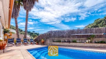 Piscina privada al aire libre en Villa Valeria con agua azul, bordes de azulejos, tumbonas y altas palmeras bajo un cielo azul nublado.