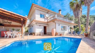 Vista exterior de Villa Valeria que cuenta con una piscina con mosaico de azulejos, una villa blanca de dos plantas con detalles en piedra, un patio cubierto con sillas blancas y altas palmeras.