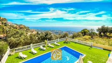 Piscina privada y césped verde en Villa Sunrise, con tumbonas blancas y una terraza con balaustrada blanca que ofrece vistas al mar y a las colinas bajo un cielo azul.