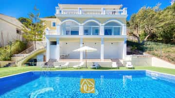 Exterior de Villa Sunrise que muestra una piscina rectangular de azulejos azules, tumbonas blancas y una villa amarilla de varias plantas con puertas de cristal arqueadas bajo un cielo despejado.