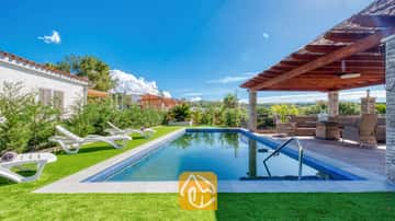 Piscina exterior y patio cubierto en Villa Palmera, que cuenta con tumbonas de plástico blanco sobre césped artificial y una pérgola de madera con asientos de mimbre.