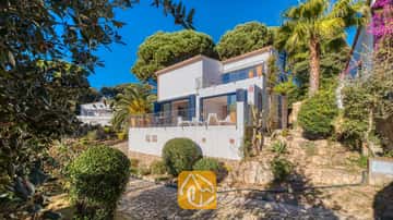 Exterior de Casa AdoRa que muestra una fachada blanca con contraventanas azules, un camino de piedra pavimentado, palmeras y pinos verdes bajo un cielo azul despejado.