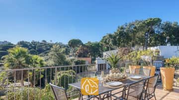 Soleada terraza en Casa AdoRa con un juego de comedor de metal equipado con platos blancos y copas, con vistas a frondosos árboles bajo un cielo azul.