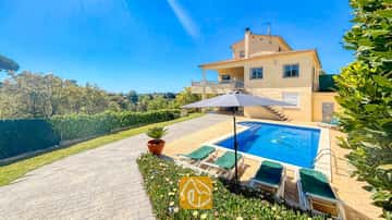 Vista exterior de Villa Holiday que muestra la casa amarilla de dos plantas, la piscina con mosaico de delfines y las hamacas verdes bajo una sombrilla gris.