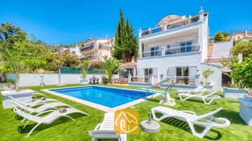 Vista exterior de la Villa Maribel que muestra una piscina rectangular, trampolín y tumbonas blancas dispuestas sobre césped artificial verde bajo un cielo azul brillante.