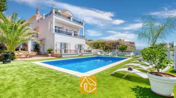 Piscina privada y tumbonas blancas sobre un césped verde en Villa Maribel, España, que cuenta con una villa blanca de varias plantas con balcones y palmeras.