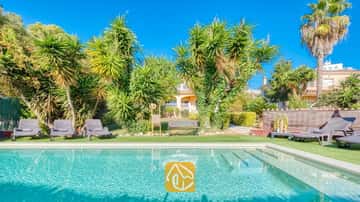 Piscina exterior con escalones integrados en Villa Summertime, rodeada de césped artificial, múltiples tumbonas y palmeras tropicales bajo un cielo azul.
