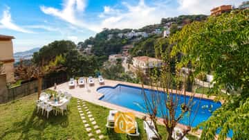 Villa Marysol cuenta con una piscina al aire libre rodeada de tumbonas blancas, una mesa de comedor en el césped y un pueblo en la ladera de la colina como telón de fondo.