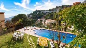 Villa Marysol cuenta con una piscina al aire libre rodeada de tumbonas blancas, una mesa de comedor en el césped y un pueblo en la ladera de la colina como telón de fondo.