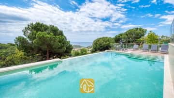 Piscina privada en Villa BellaVista con azulejos color turquesa, una terraza con barandillas de cristal equipada con tumbonas grises y vistas al mar y a los pinos.