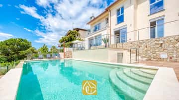 Gran piscina al aire libre con escalones curvos en Villa BellaVista. El exterior revestido en piedra incluye terrazas con barandillas de cristal, suelos de madera y tumbonas grises rodeadas de frondosos árboles.