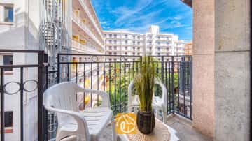 Balcón del Apartamento Gaudí con dos sillas de plástico blancas, una pequeña mesa con una planta en maceta y una barandilla de metal con vistas a los edificios blancos colindantes.