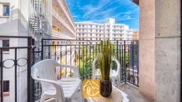 Balcón del Apartamento Gaudí con dos sillas de plástico blancas, una pequeña mesa con una planta en maceta y una barandilla de metal con vistas a los edificios blancos colindantes.