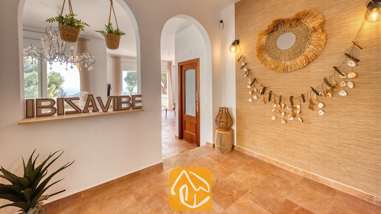 Pasillo interior de Villa Ibiza con aberturas en arco, una pared de acento de paja texturizada decorada con una guirnalda de conchas y un espejo de paja, y suelos de baldosa.