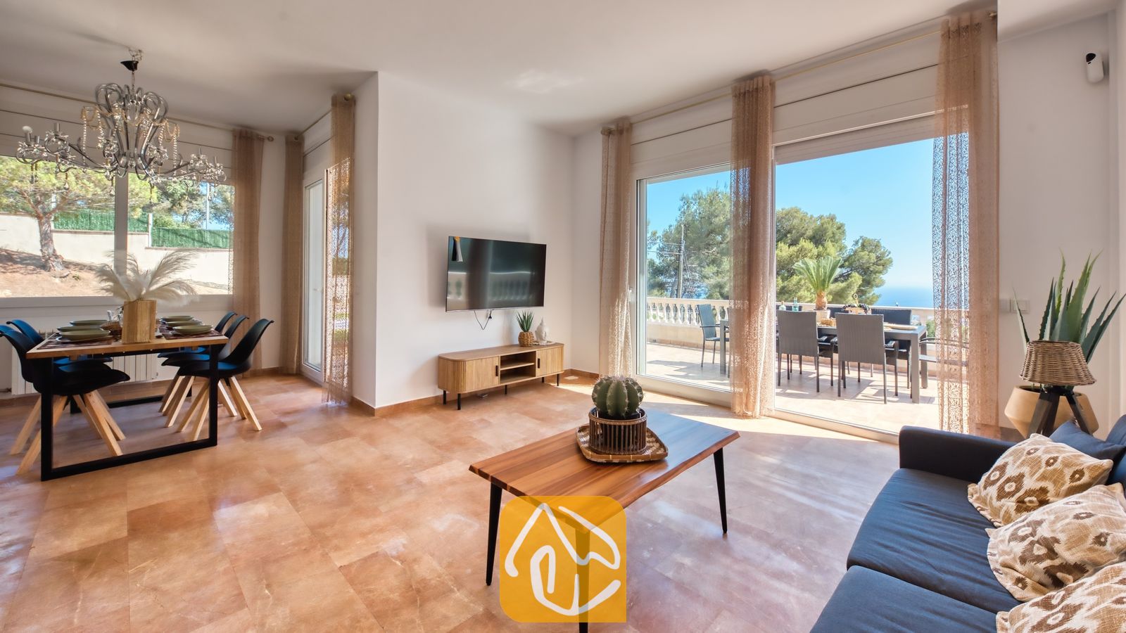 Espacioso salón y comedor en Villa Ibiza que cuenta con un sofá oscuro, mesa de centro de madera con un cactus en maceta, televisión montada en la pared y grandes puertas correderas de cristal que se abren a una soleada terraza con