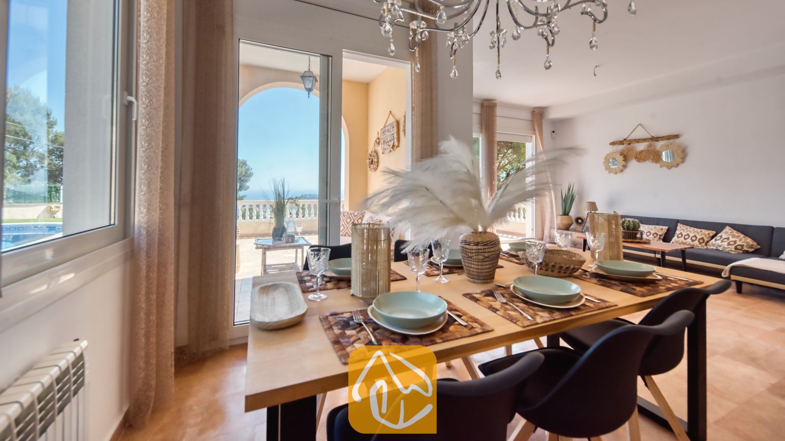 Juego de comedor de madera con servicio de mesa, cristalería y un gran centro de mesa de plumas, con puertas de cristal que se abren a una terraza soleada en Villa Ibiza.