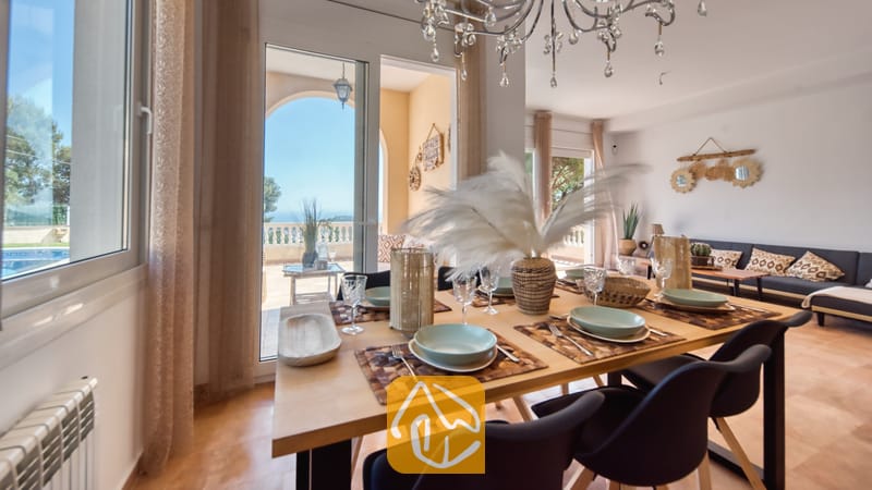 Juego de comedor de madera con servicio de mesa, cristalería y un gran centro de mesa de plumas, con puertas de cristal que se abren a una terraza soleada en Villa Ibiza.