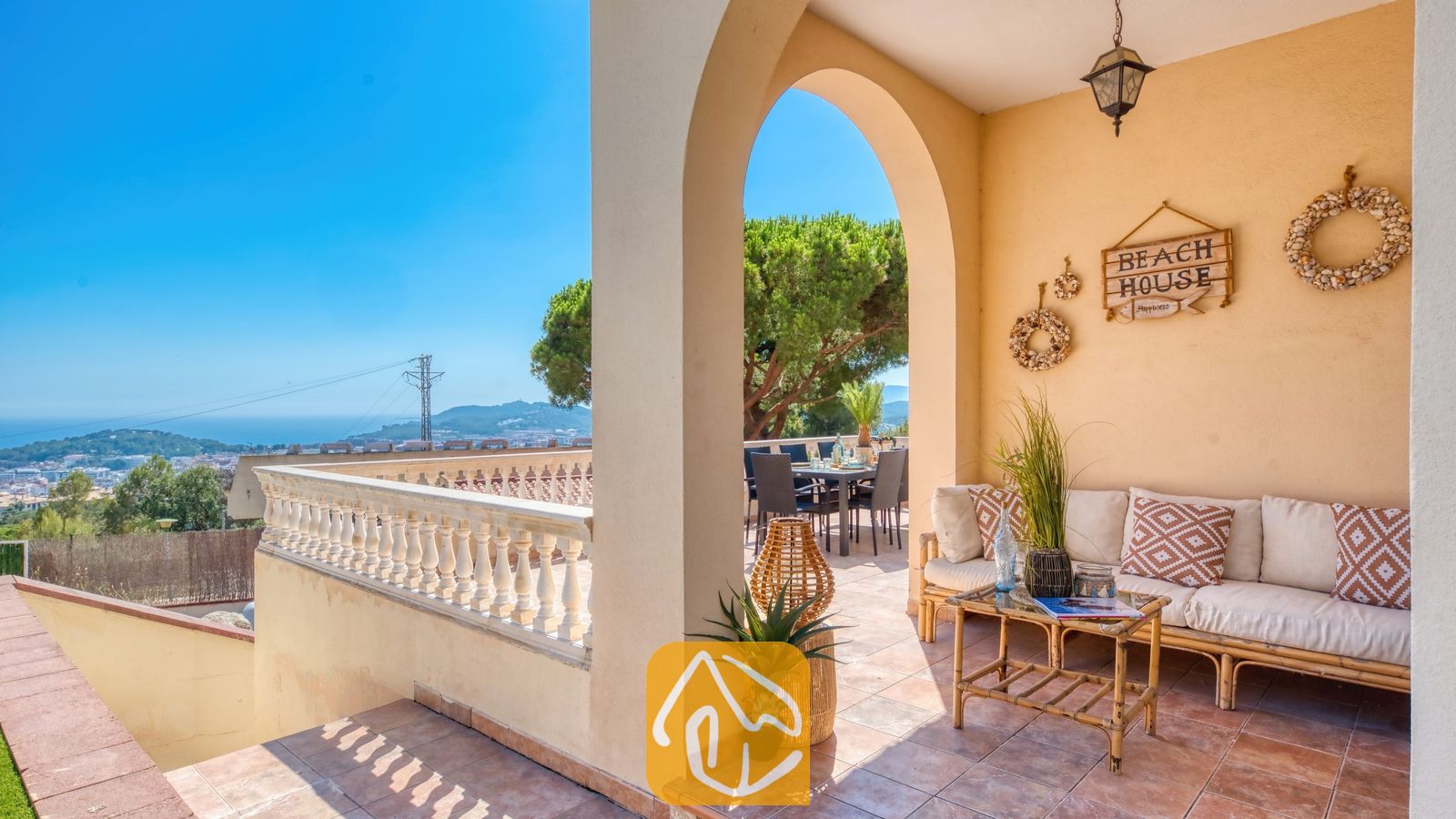 Terraza cubierta en Villa Ibiza con un sofá beige, cojines estampados, una mesa de centro de madera y un arco que enmarca una zona de asientos al aire libre y una vista lejana al paisaje.