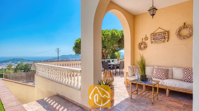 Terraza cubierta en Villa Ibiza con un sofá beige, cojines estampados, una mesa de centro de madera y un arco que enmarca una zona de asientos al aire libre y una vista lejana al paisaje.