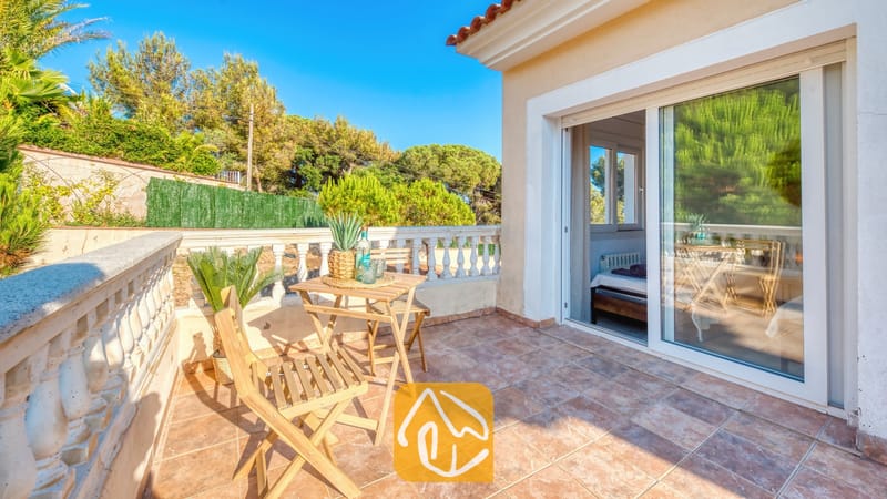 Soleada terraza en Villa Ibiza equipada con una mesa de madera y dos sillas plegables, suelo de baldosas, barandilla de balaustrada y puertas corredizas de cristal que dejan ver el interior de un dormitorio.