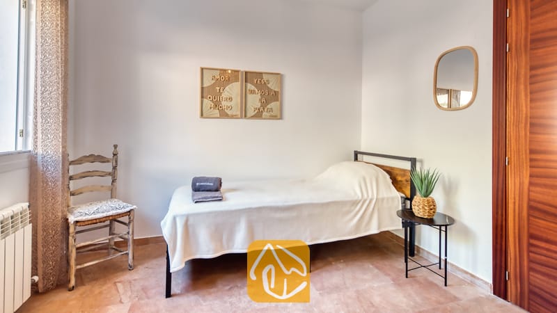 Cama individual con sábanas blancas y una toalla gris doblada junto a una mesita auxiliar negra y una silla de madera rústica en Villa Ibiza.