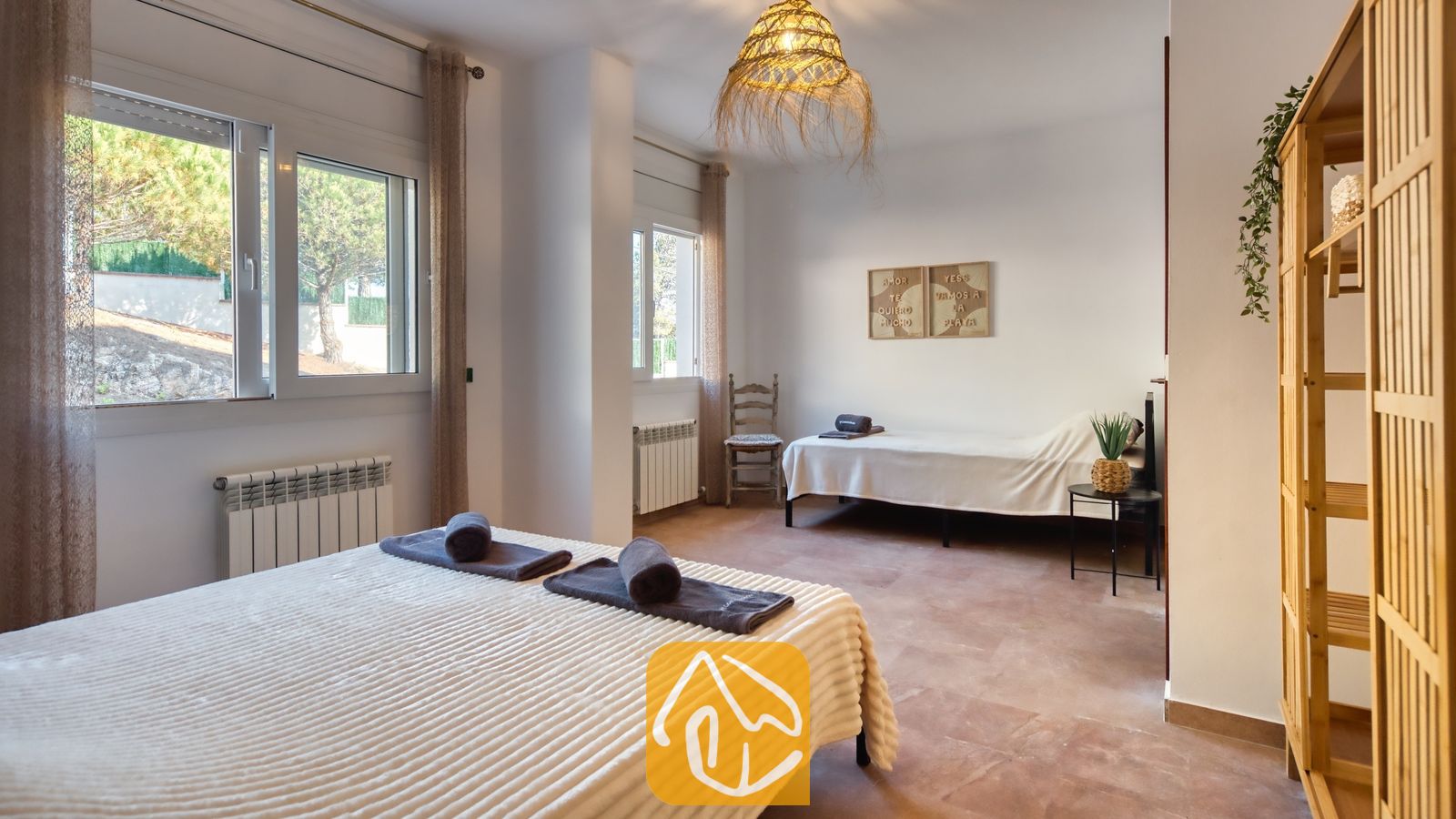 Dormitorio con dos camas individuales, toallas oscuras enrolladas, paredes blancas y una lámpara colgante de mimbre trenzado del techo.