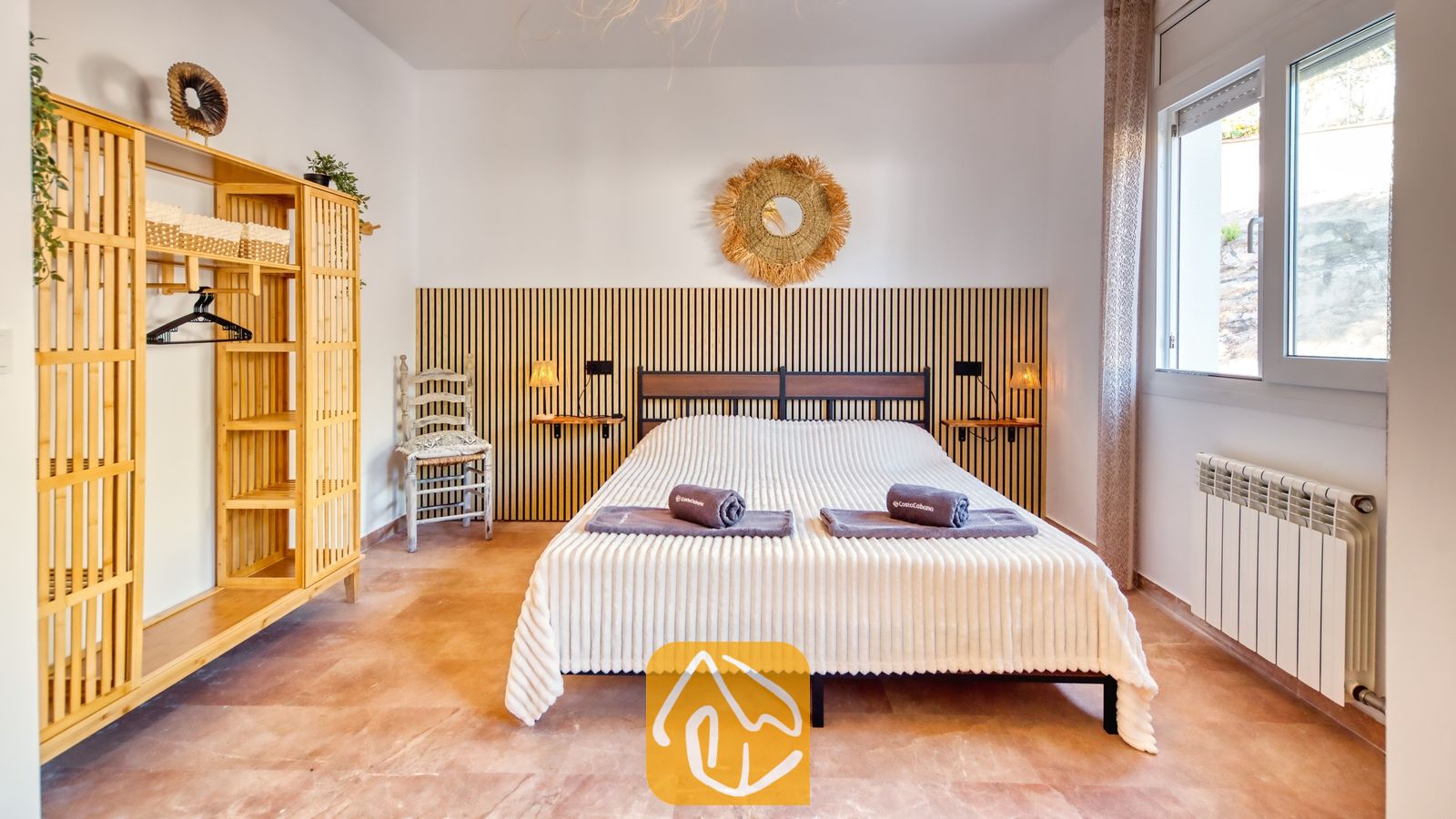 Dormitorio con una cama doble que incluye toallas enrolladas, cabecero de listones de madera, armario de bambú y suelo de baldosas.