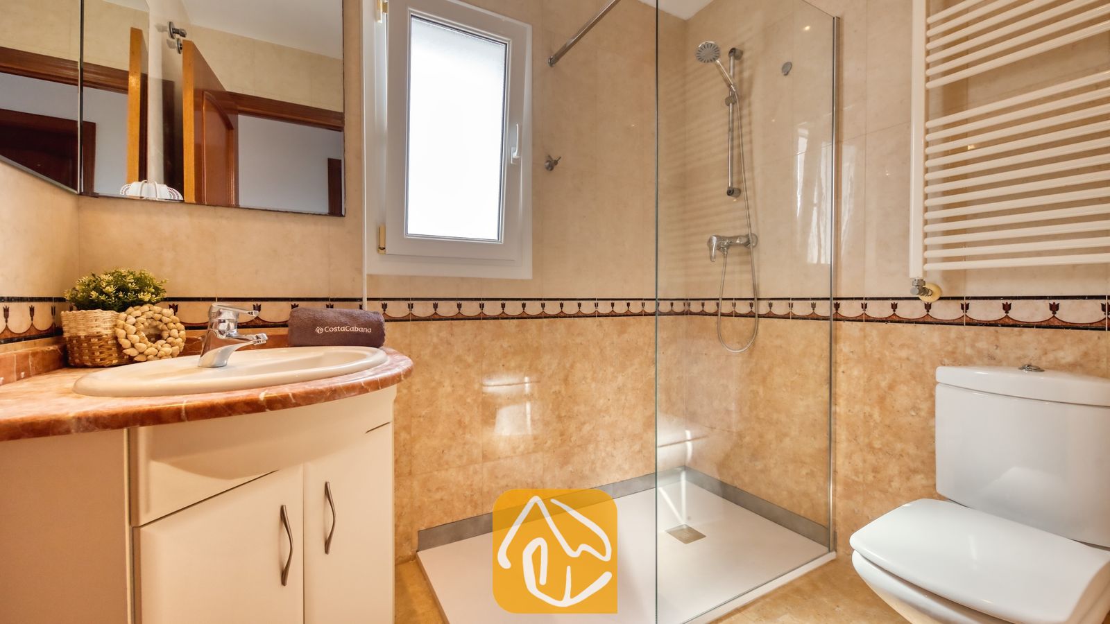 Baño moderno con paredes revestidas de azulejos en tonos beige, ducha acristalada a ras de suelo, mueble de baño blanco con lavabo de forma redondeada e inodoro blanco.