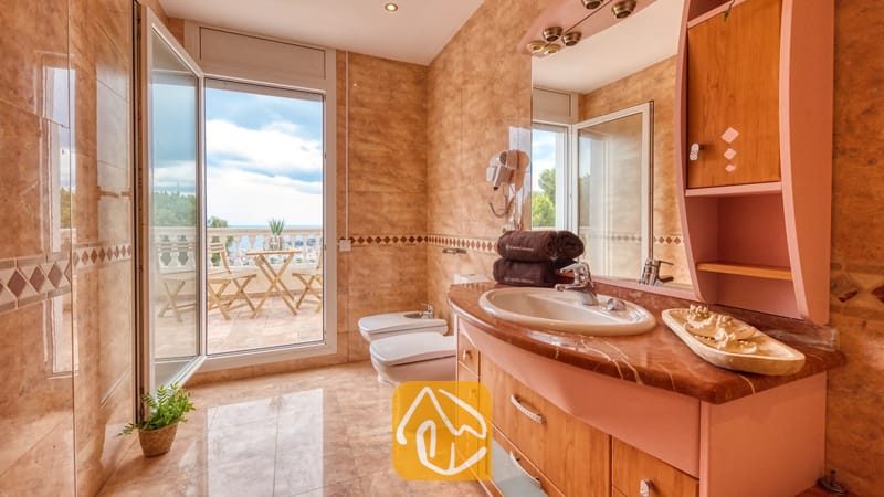 Baño con paredes de azulejos de efecto mármol, un mueble de madera con lavabo integrado y una puerta de cristal abierta que da a un balcón con vistas al mar.