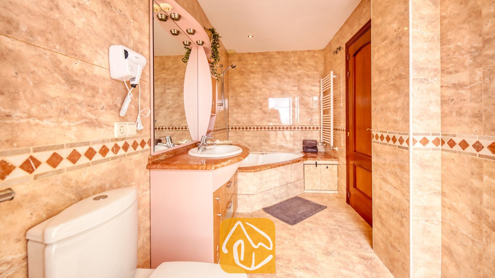 Baño equipado con azulejos con patrón de mármol en tonos beige, una bañera esquinera y un tocador con un espejo arqueado de color rosa.