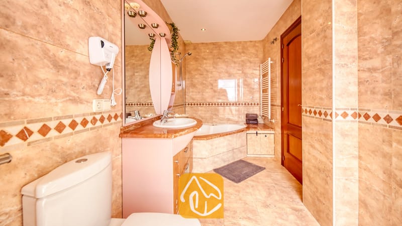 Baño equipado con azulejos con patrón de mármol en tonos beige, una bañera esquinera y un tocador con un espejo arqueado de color rosa.