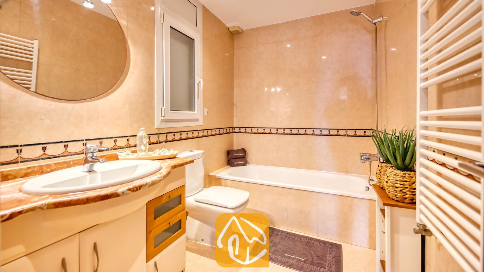 Baño con azulejos de pared con efecto de mármol en tonos beige, una bañera de cerámica blanca, un lavabo con tocador y espejo redondo, y una planta en maceta.