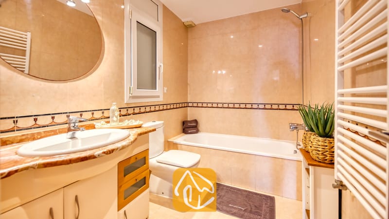Baño con azulejos de pared con efecto de mármol en tonos beige, una bañera de cerámica blanca, un lavabo con tocador y espejo redondo, y una planta en maceta.