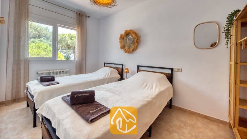 Dormitorio con dos camas individuales con estructura de metal oscuro, ropa de cama blanca, toallas grises dobladas y una ventana con vistas a los árboles.