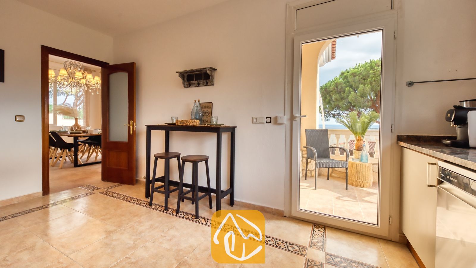 Vista interior de Villa Ibiza que muestra una zona de cocina con una mesa consola alta, dos taburetes de madera, suelos de baldosa y una puerta de cristal abierta que da a un balcón.