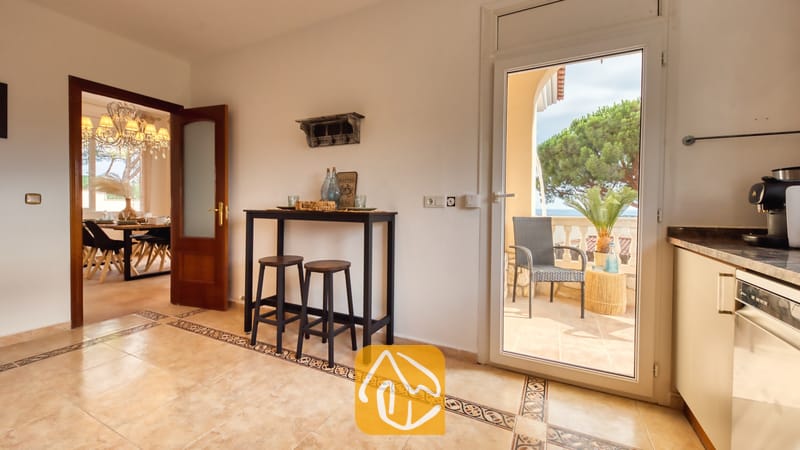 Vista interior de Villa Ibiza que muestra una zona de cocina con una mesa consola alta, dos taburetes de madera, suelos de baldosa y una puerta de cristal abierta que da a un balcón.