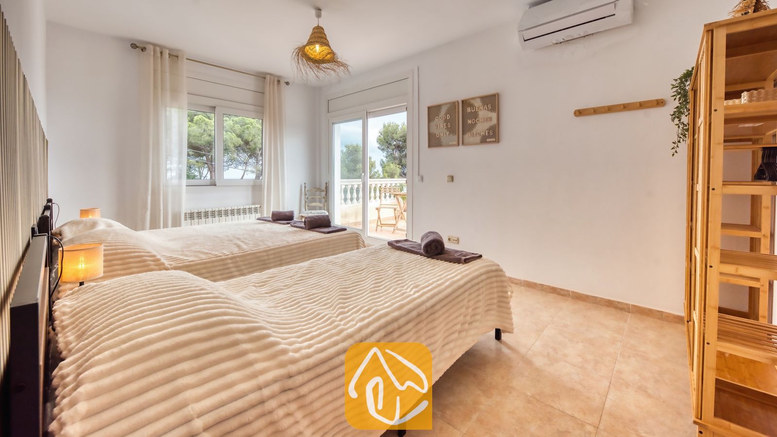 Dormitorio con dos camas individuales que incluye mantas texturizadas en tono beige, una estantería de madera y una puerta de cristal con acceso a un balcón desde el que se divisan árboles verdes.