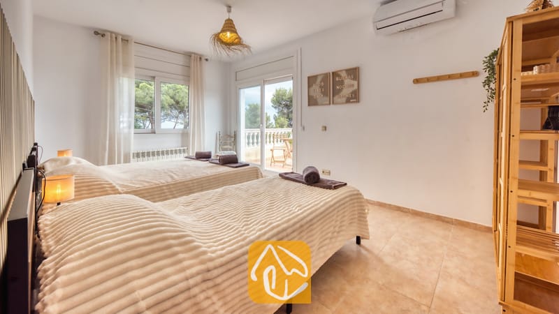 Dormitorio con dos camas individuales que incluye mantas texturizadas en tono beige, una estantería de madera y una puerta de cristal con acceso a un balcón desde el que se divisan árboles verdes.
